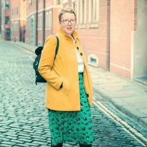 A woman stands in the street wearing a yellow coat, green skirt and a backpack