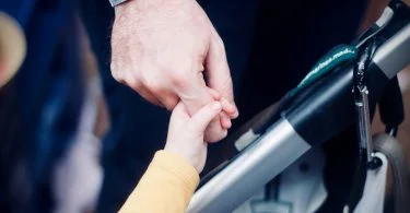 A child's hand holds the index finger of a man. In between the two is the handle of a pushchair