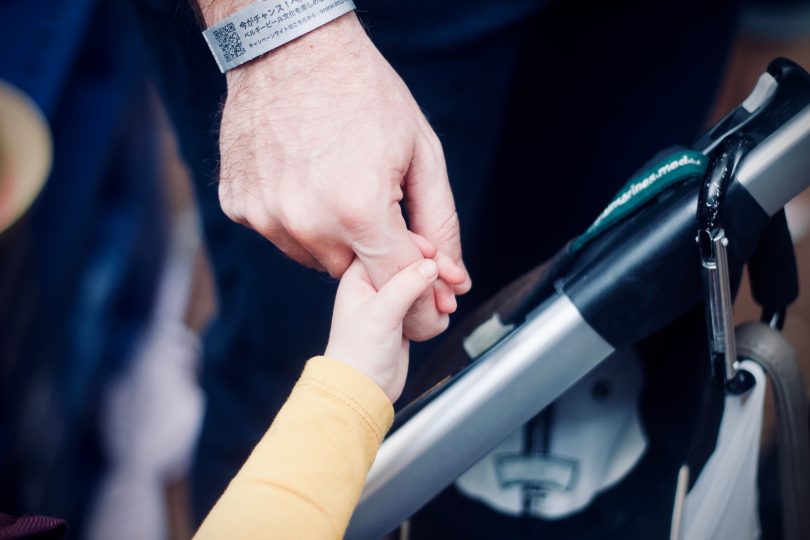 A child's hand holds the index finger of a man. In between the two is the handle of a pushchair